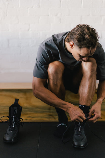 A man in a dark gray t-shirt and black athletic socks is crouching down to tie the laces on his black running shoe. Another black running shoe lies on the floor to his left, set against a white brick wall and wooden floor background.