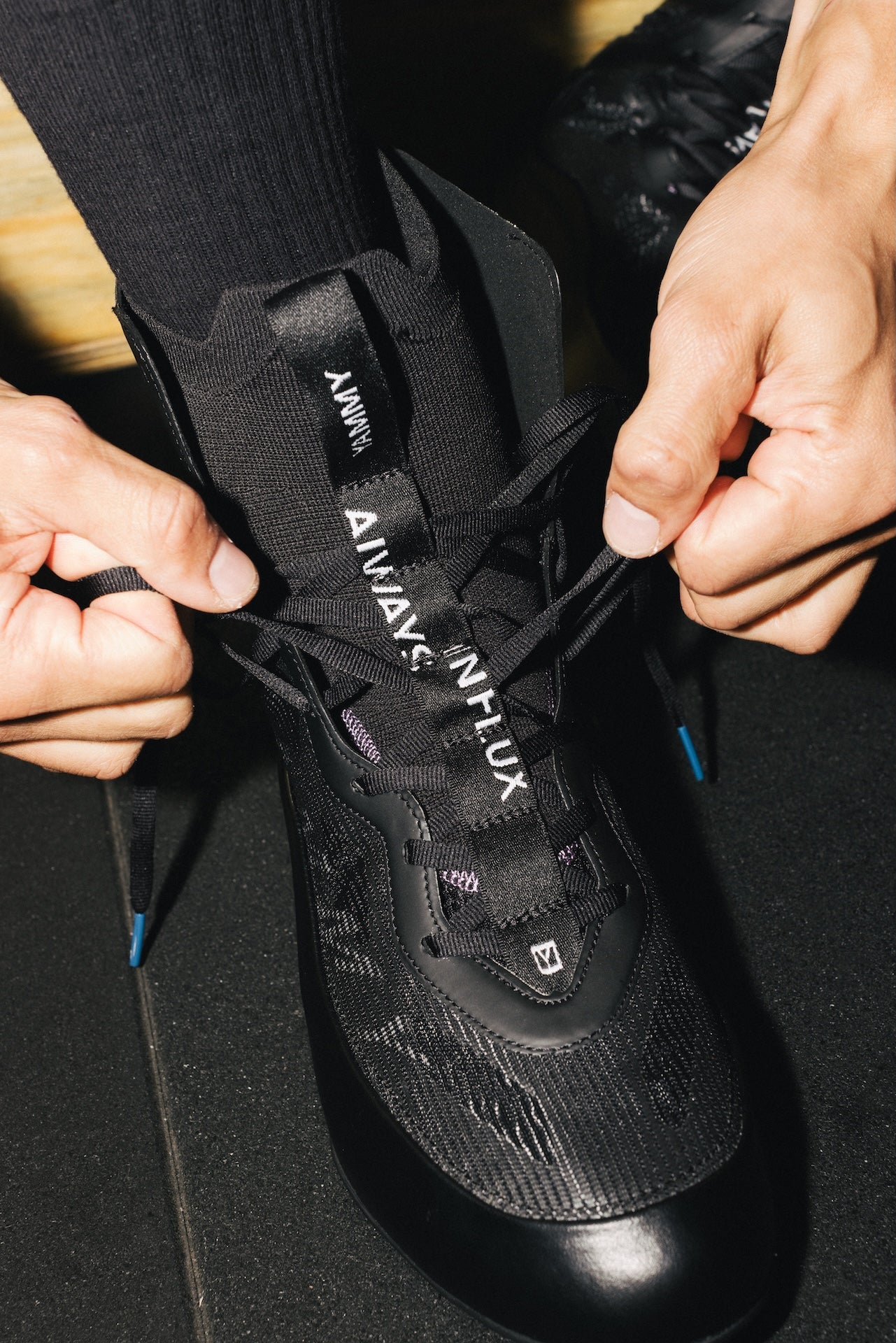 A close-up of a person's hands tying the black laces of a black high-top sneaker. The shoe features a textured mesh upper, a glossy black toe cap, and a black tongue strap with the white text 'ALWAYS INFLUX'.