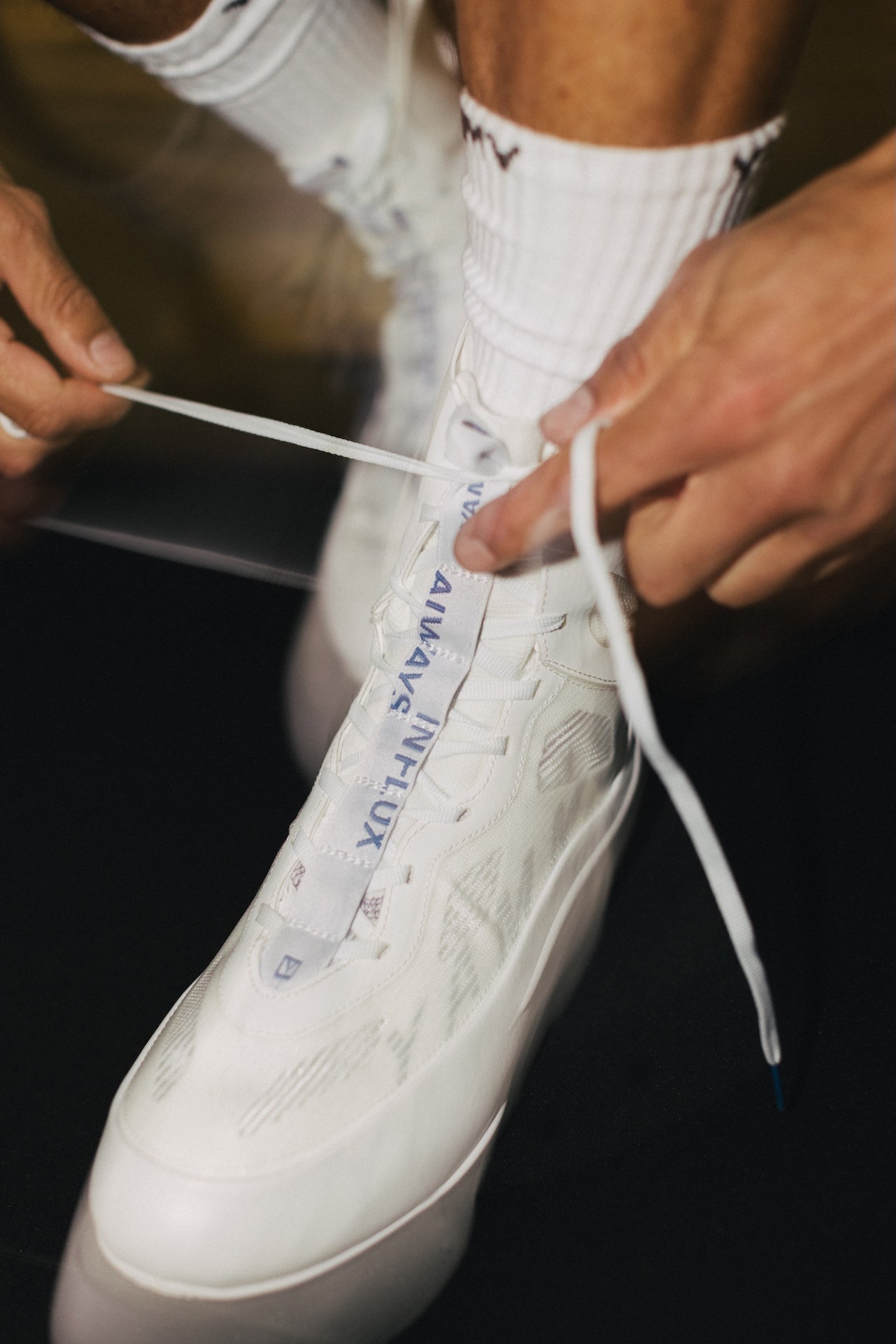 A close-up of a person tying the laces of a white high-top sneaker. The shoe has visible branding text reading 'AIRAVALUX' on the tongue area, and the person is wearing white crew socks against a dark background.