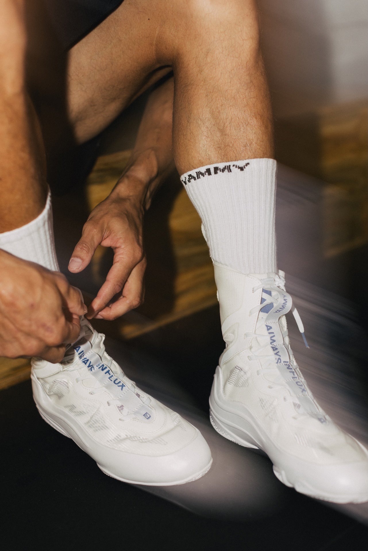 A close-up lifestyle shot of a person sitting down, tying the laces of a pair of white high-top sneakers. The person is wearing white crew socks with a black 'RAMMY' logo.