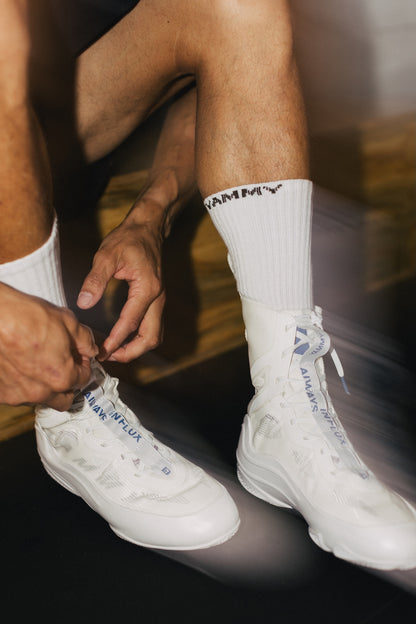 A close-up lifestyle shot of a person sitting down, tying the laces of a pair of white high-top sneakers. The person is wearing white crew socks with a black 'RAMMY' logo.