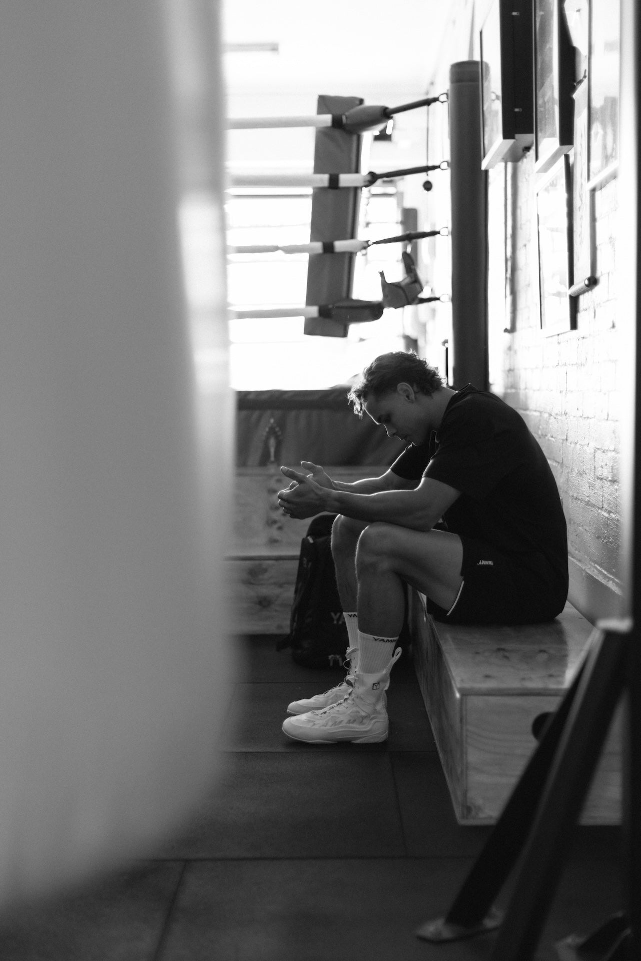 A black and white photograph of a man sitting on a wooden bench in a gym, looking down at his hands. He is wearing a dark t-shirt, shorts, and high-top sneakers.