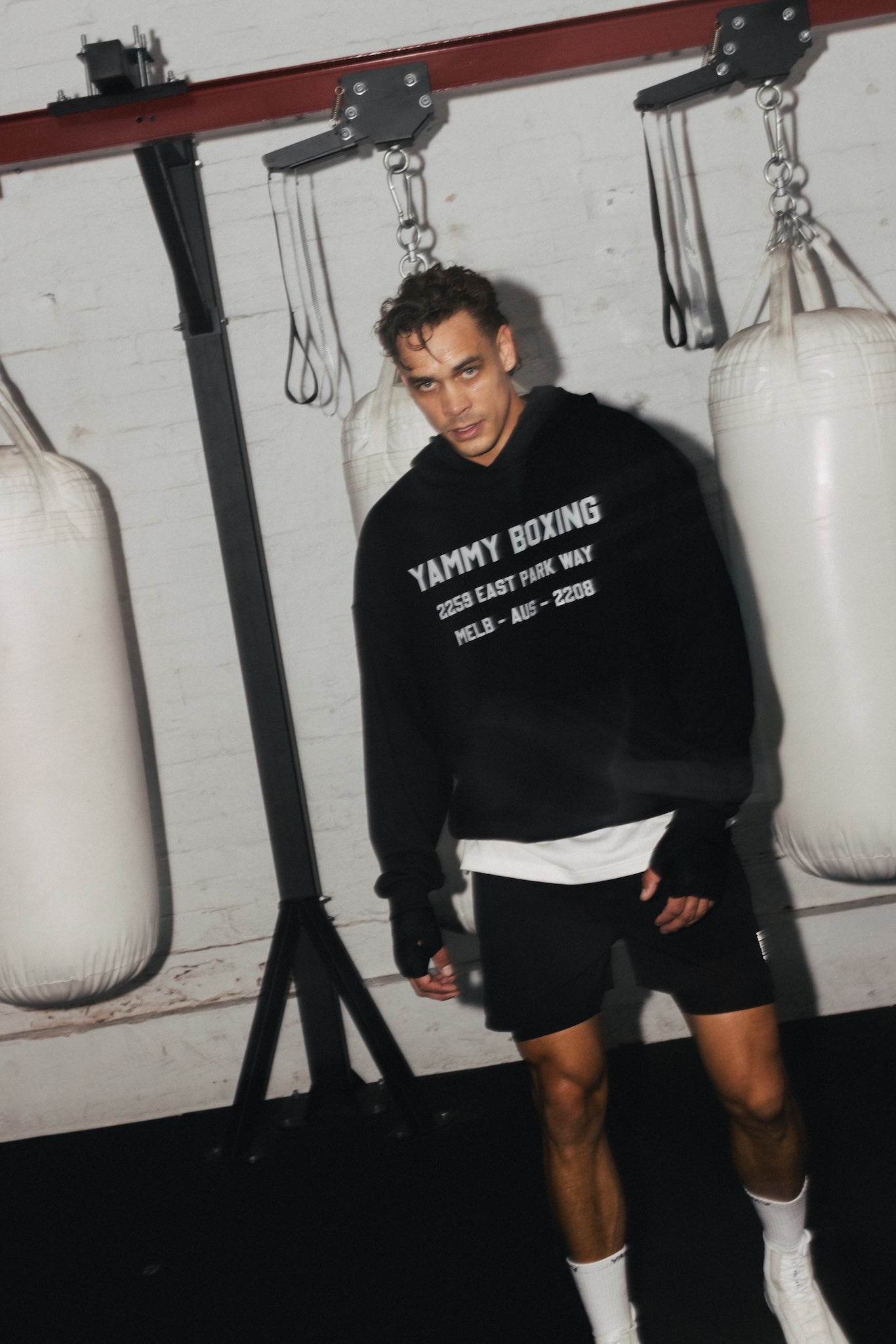 A man stands in a boxing gym wearing a black hoodie with white text that reads 'YAMMY BOXING'.