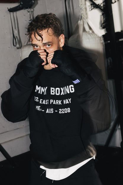 A man in a boxing stance wearing a black hoodie with 'YAMMY BOXING' printed in white text on the front, set against a dark gym background with punching bags.