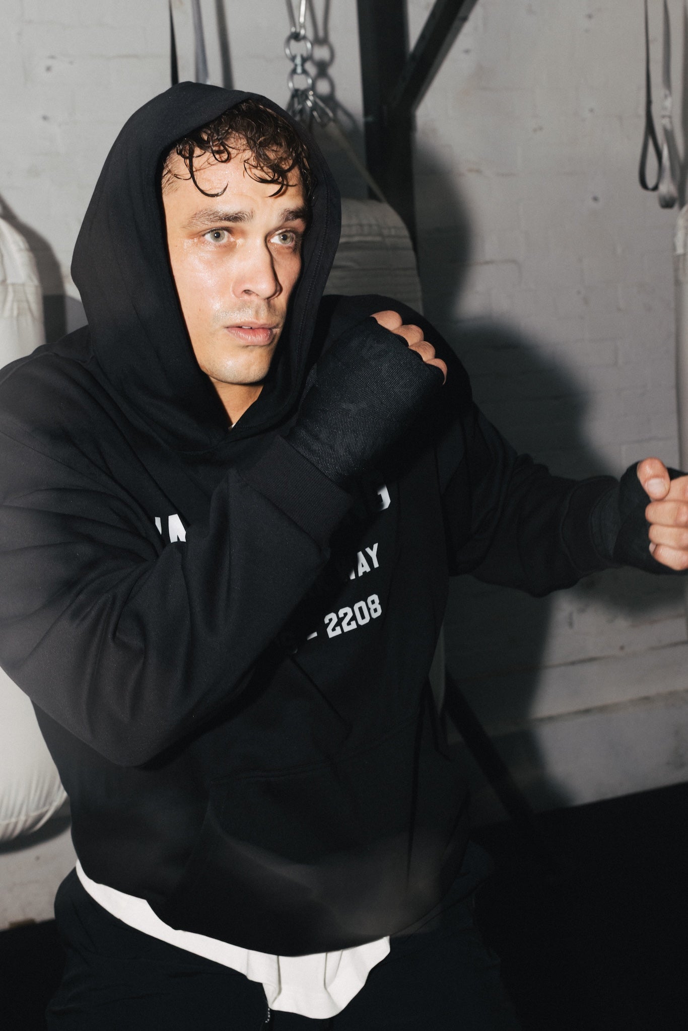 A man in a black hoodie and black hand wraps poses in a gym setting with punching bags and a brick wall in the background.