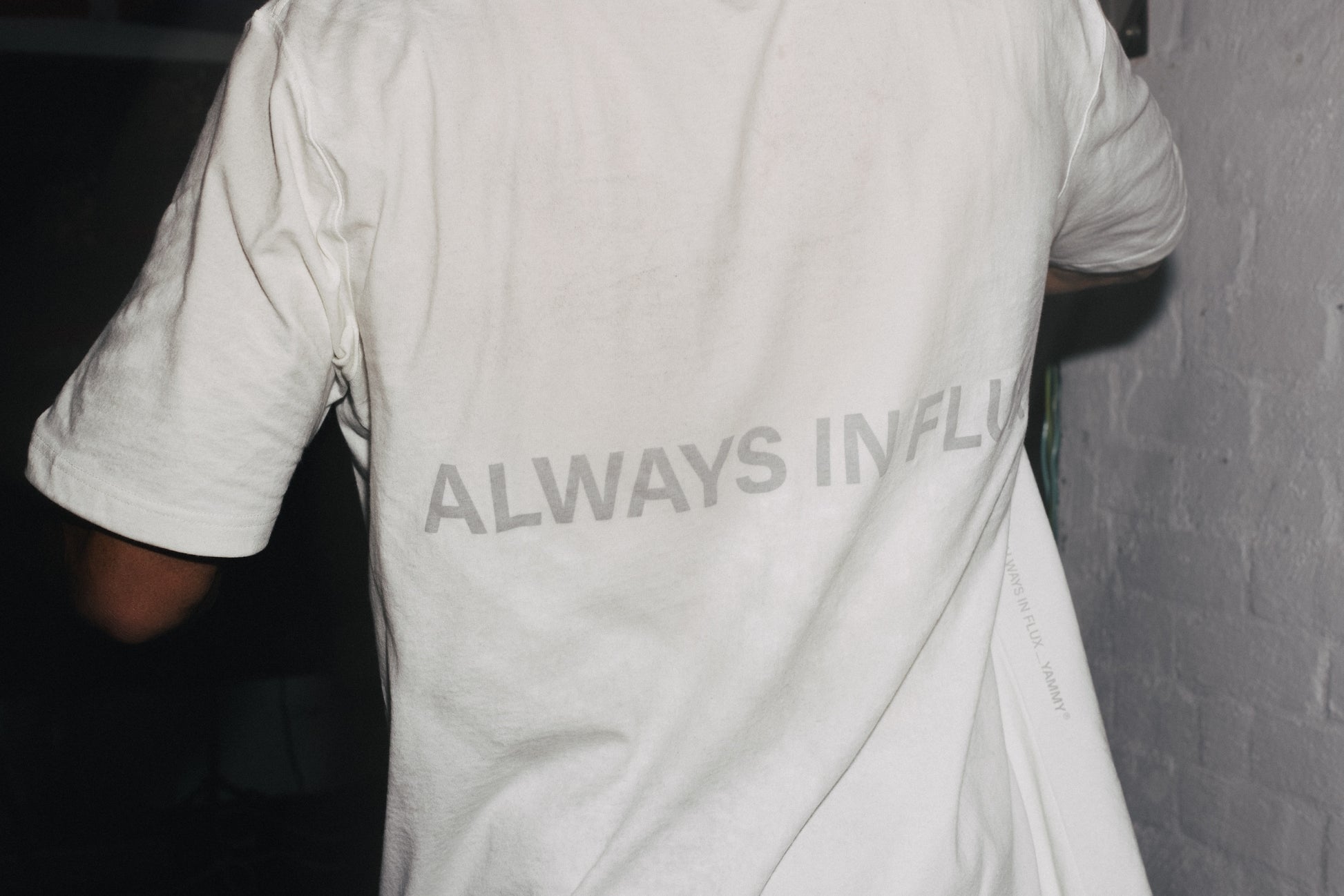 A person wearing a white t-shirt with the text 'ALWAYS IN FLUX' printed in gray across the back, standing against a white brick wall.