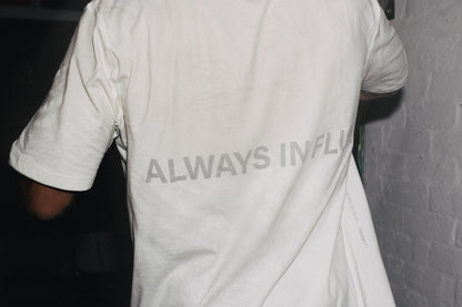 A person wearing a white t-shirt with the text 'ALWAYS IN FLUX' printed in gray across the back, standing against a white brick wall.