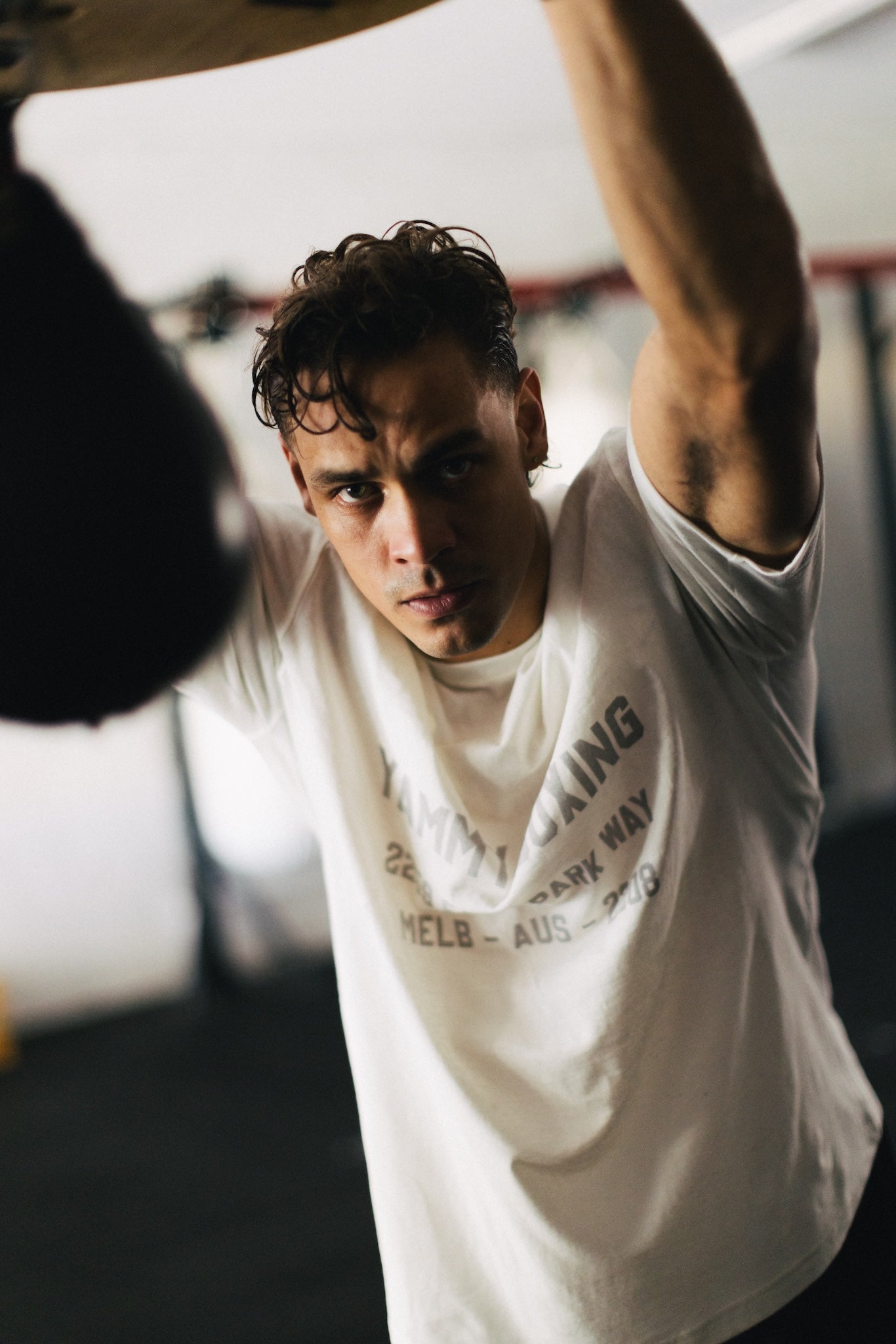 A man in a gym wearing a white graphic t-shirt with black text, captured from a low angle with one arm raised overhead.