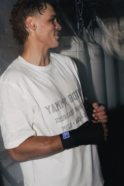 A man in a gym wearing a white graphic t-shirt and a black hand wrap, standing in front of hanging punching bags.
