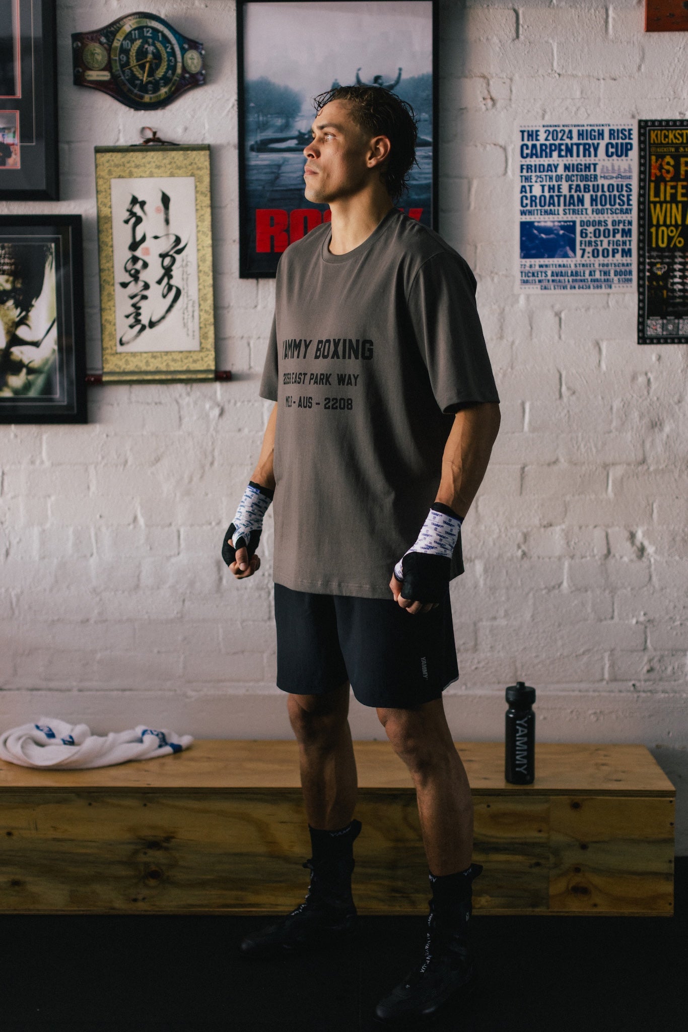 A man wearing a gray 'VINY BOXING' t-shirt, black boxing shorts, and black boxing boots stands in a gym with a white brick wall background, looking upward.