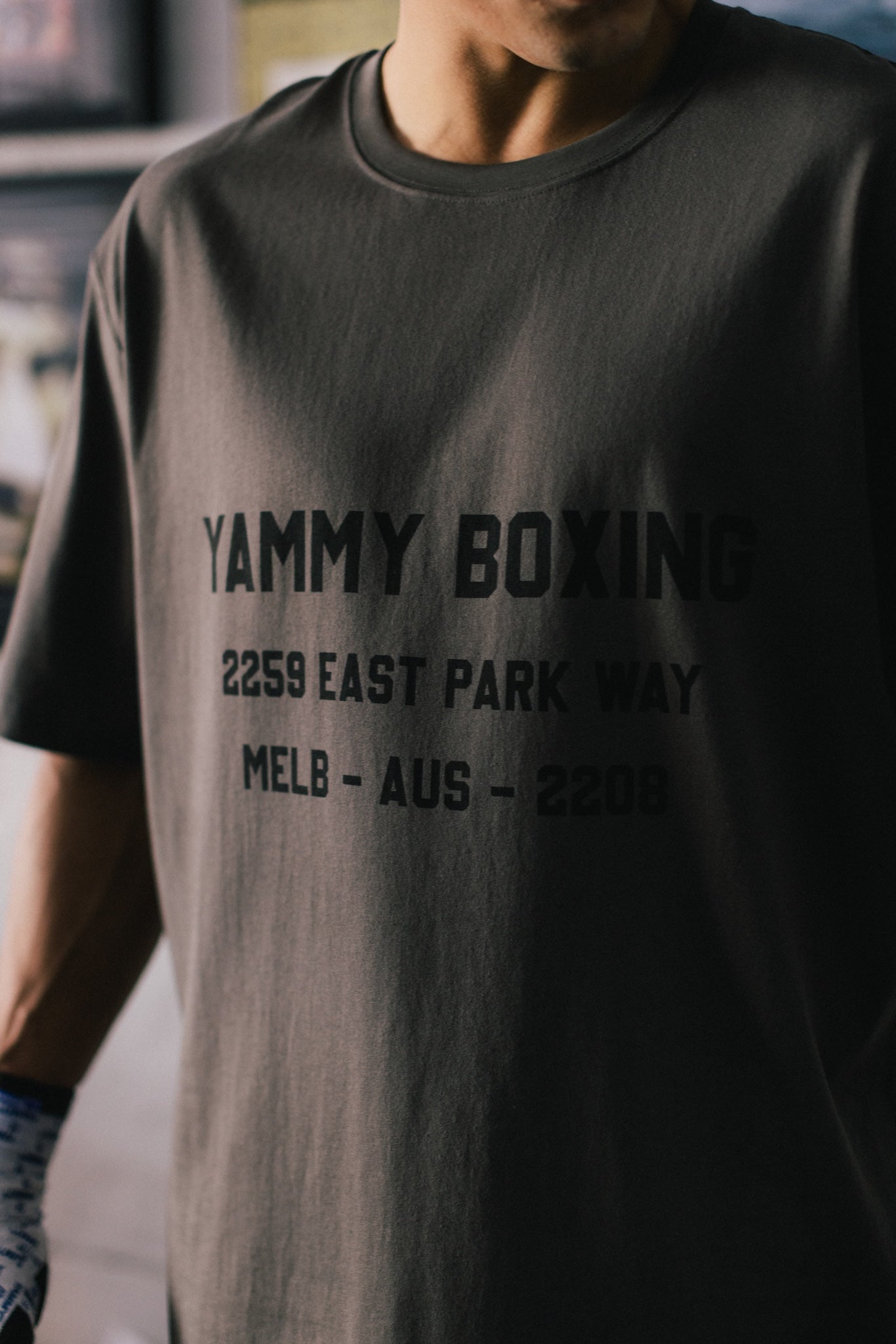 A close-up of a person wearing a dark charcoal gray t-shirt with black printed text reading 'YAMMY BOXING 2259 EAST PARK WAY MELB - AUS - 2208'.