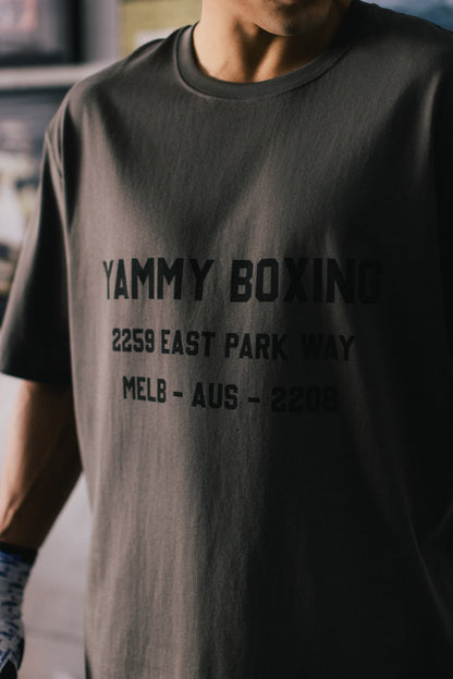 A close-up of a person wearing a dark charcoal gray t-shirt with black printed text reading 'YAMMY BOXING 2259 EAST PARK WAY MELB - AUS - 2208'.