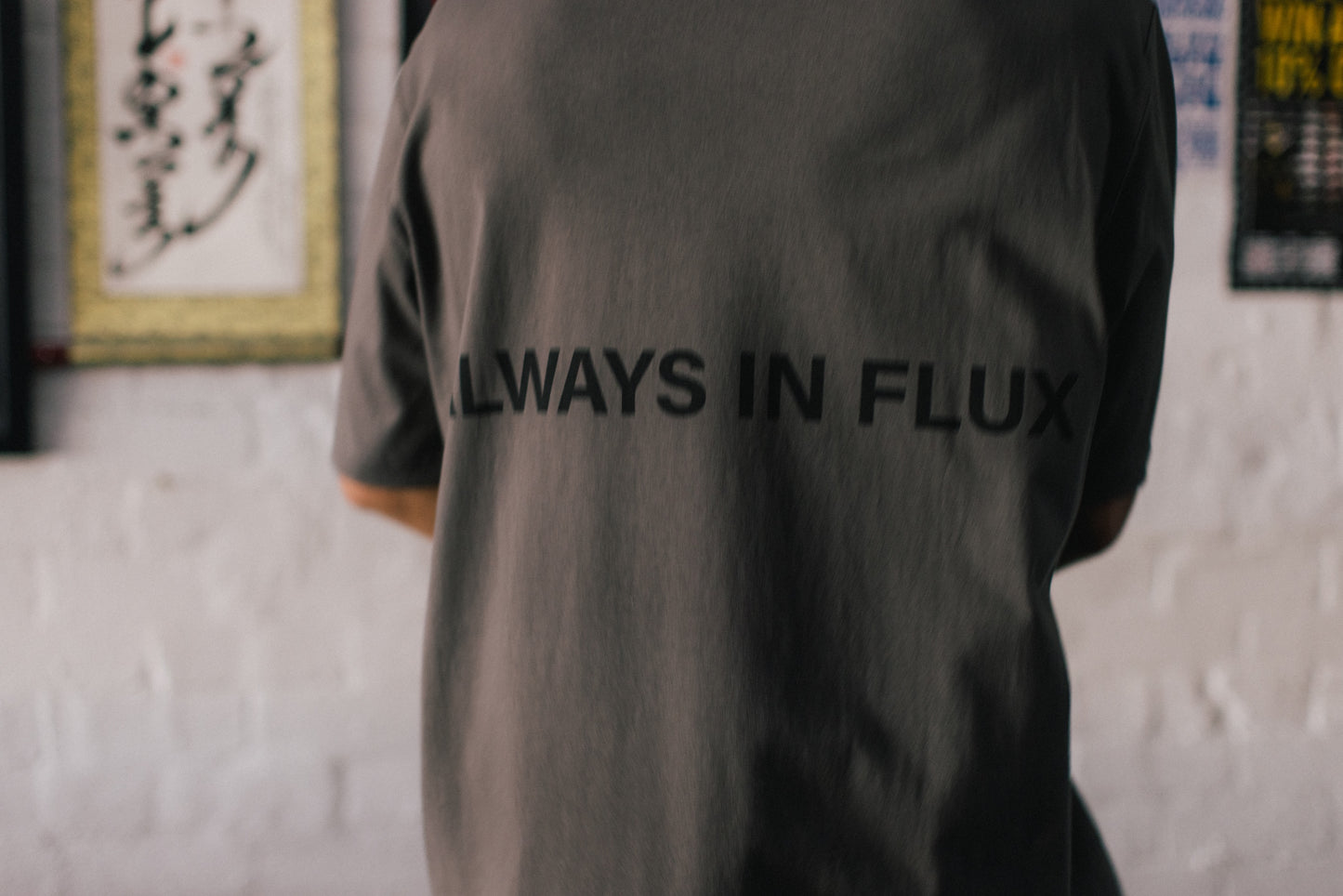 A person viewed from behind wearing a dark gray oversized T-shirt with the words 'ALWAYS IN FLUX' printed in black across the back, standing against a white brick wall.