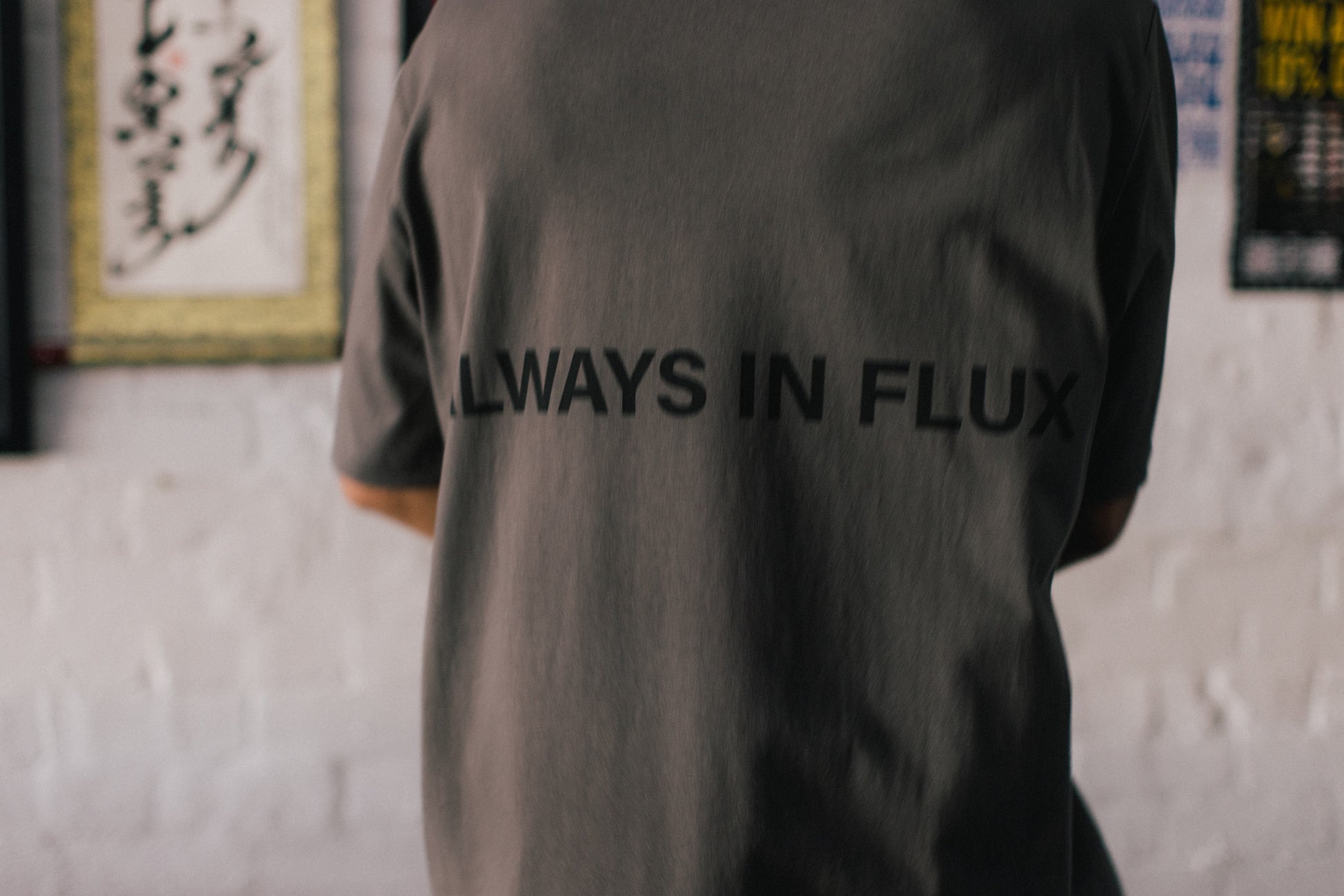 A person viewed from behind wearing a dark gray oversized T-shirt with the words 'ALWAYS IN FLUX' printed in black across the back, standing against a white brick wall.