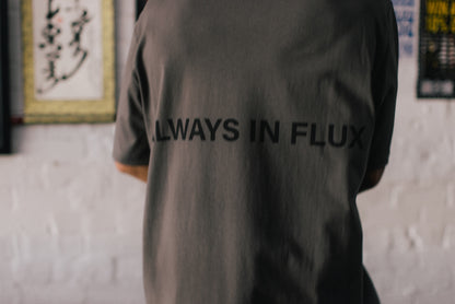 A person viewed from behind wearing a dark gray oversized T-shirt with the words 'ALWAYS IN FLUX' printed in black across the back, standing against a white brick wall.