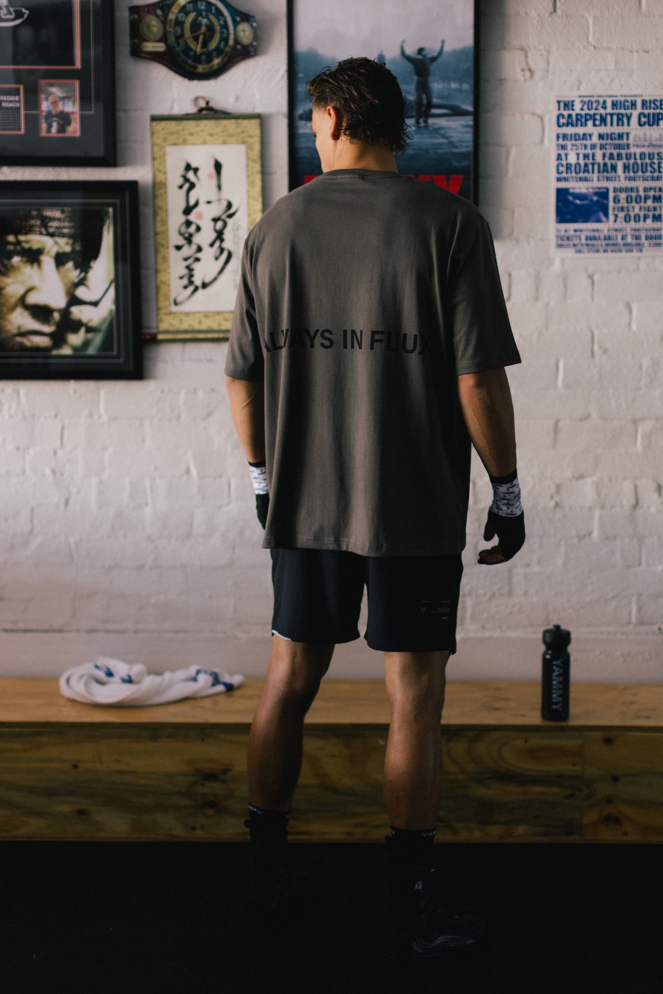 A man stands with his back to the camera wearing a dark gray t-shirt with 'ALWAYS IN FLUX' printed on the back, black athletic shorts, and gloves in a gym setting with a white brick wall and framed posters behind him.