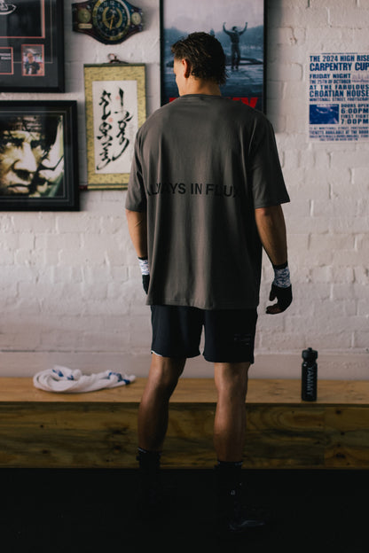 A man stands with his back to the camera wearing a dark gray t-shirt with 'ALWAYS IN FLUX' printed on the back, black athletic shorts, and gloves in a gym setting with a white brick wall and framed posters behind him.