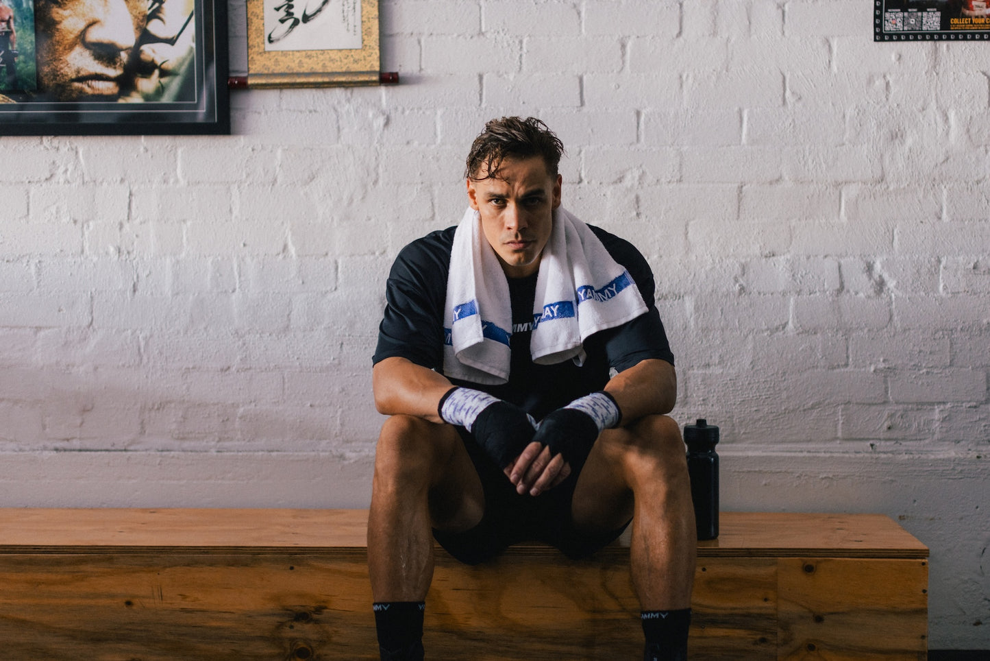 A male athlete sits on a wooden bench against a white brick wall, wearing a black athletic shirt, black shorts, black hand wraps, and a white towel with blue branding around his neck.