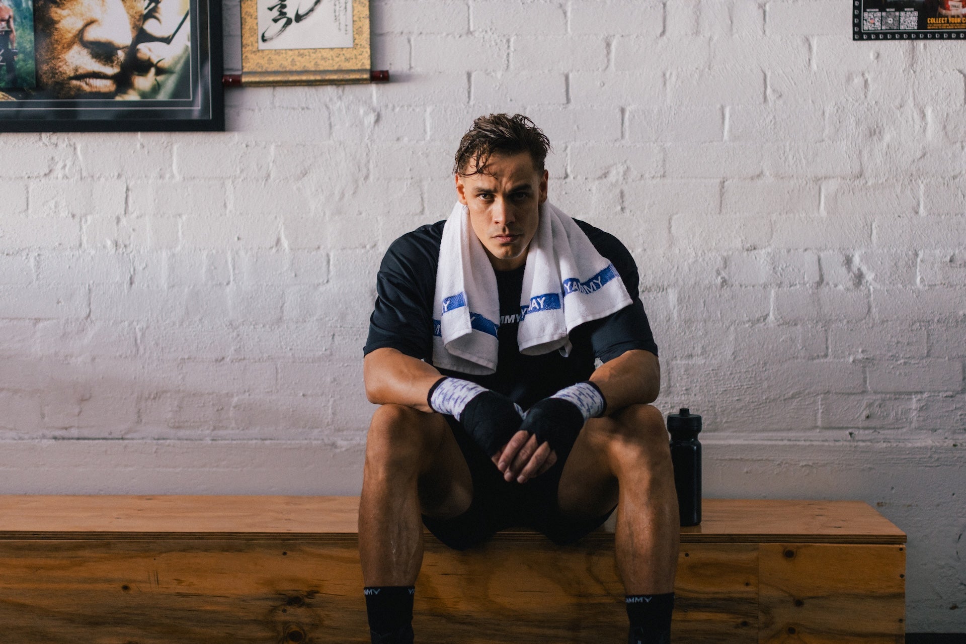 A male athlete sits on a wooden bench against a white brick wall, wearing a black athletic shirt, black shorts, black hand wraps, and a white towel with blue branding around his neck.