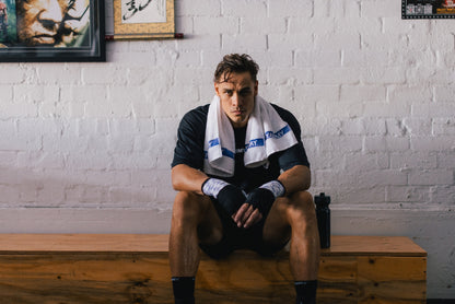 A male athlete sits on a wooden bench against a white brick wall, wearing a black athletic shirt, black shorts, black hand wraps, and a white towel with blue branding around his neck.