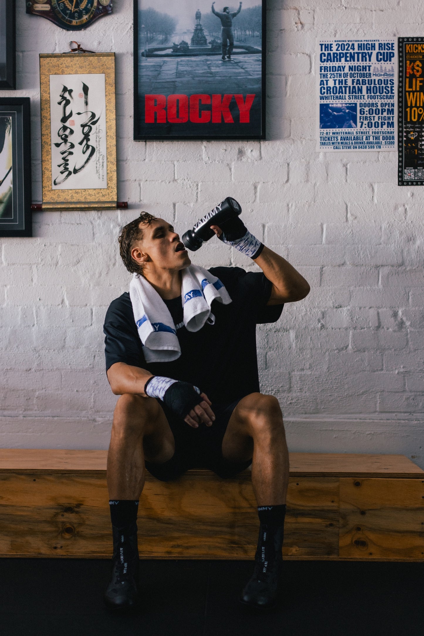 A man in a black athletic outfit and boxing gloves sits on a wooden bench in an indoor gym setting, drinking from a black bottle while wearing a white towel around his neck. The background features a white brick wall with framed posters, including a 'Rocky' poster.