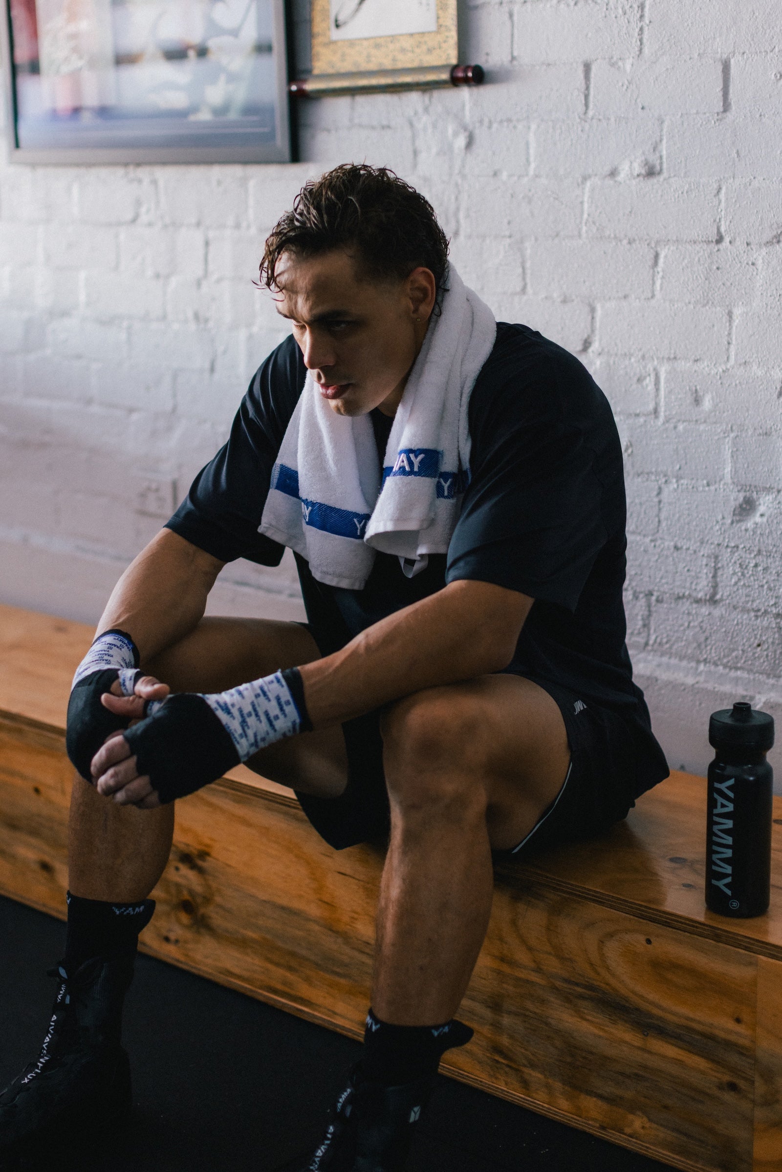 A man in athletic wear sits on a wooden bench in a gym, wrapping his hands and wearing a white towel over his shoulders. A black Yammy water bottle is on the bench next to him.