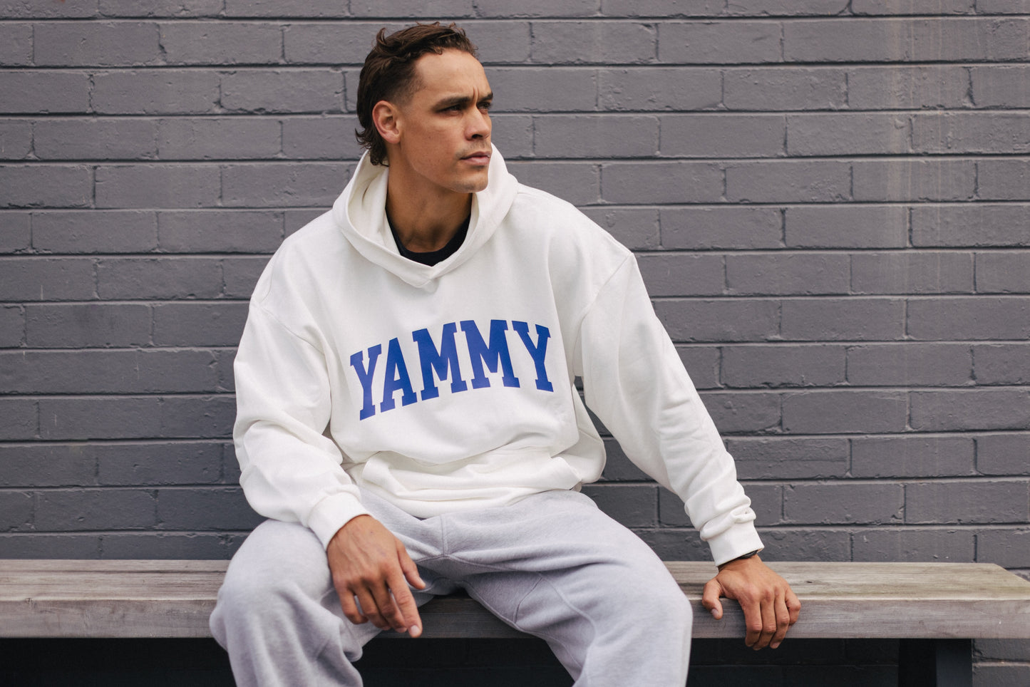 A man sitting on a wooden bench against a gray brick wall, wearing a white hoodie with 'YAMMY' printed in blue across the chest and light gray sweatpants.