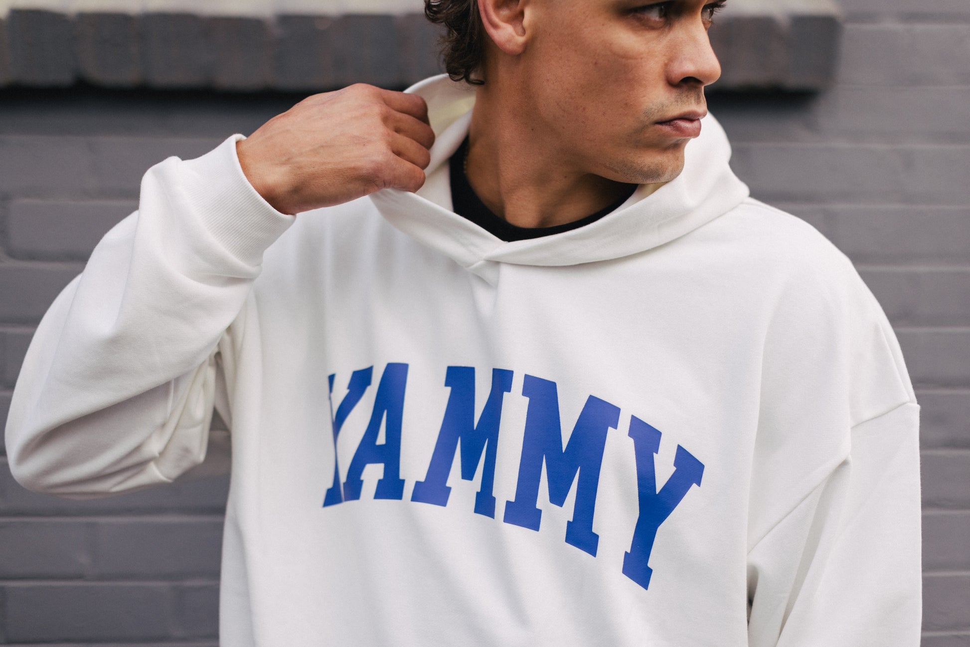 A man wearing a white oversized hoodie with 'KAMMY' printed in blue arched letters across the chest, standing against a gray brick wall.