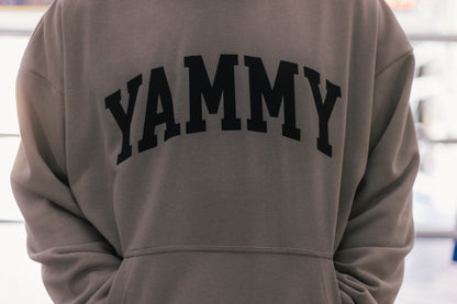 A close-up back view of a person wearing a grey hoodie with the word 'YAMMY' printed across the back in black varsity-style block letters.