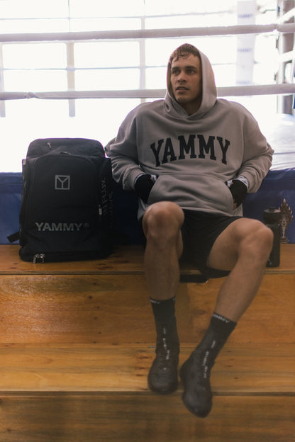 A man sits on wooden steps in a boxing ring wearing a gray hoodie with 'YAMMY' printed across the chest, black boxing shorts, and black boots. A black YAMMY backpack is resting beside him.