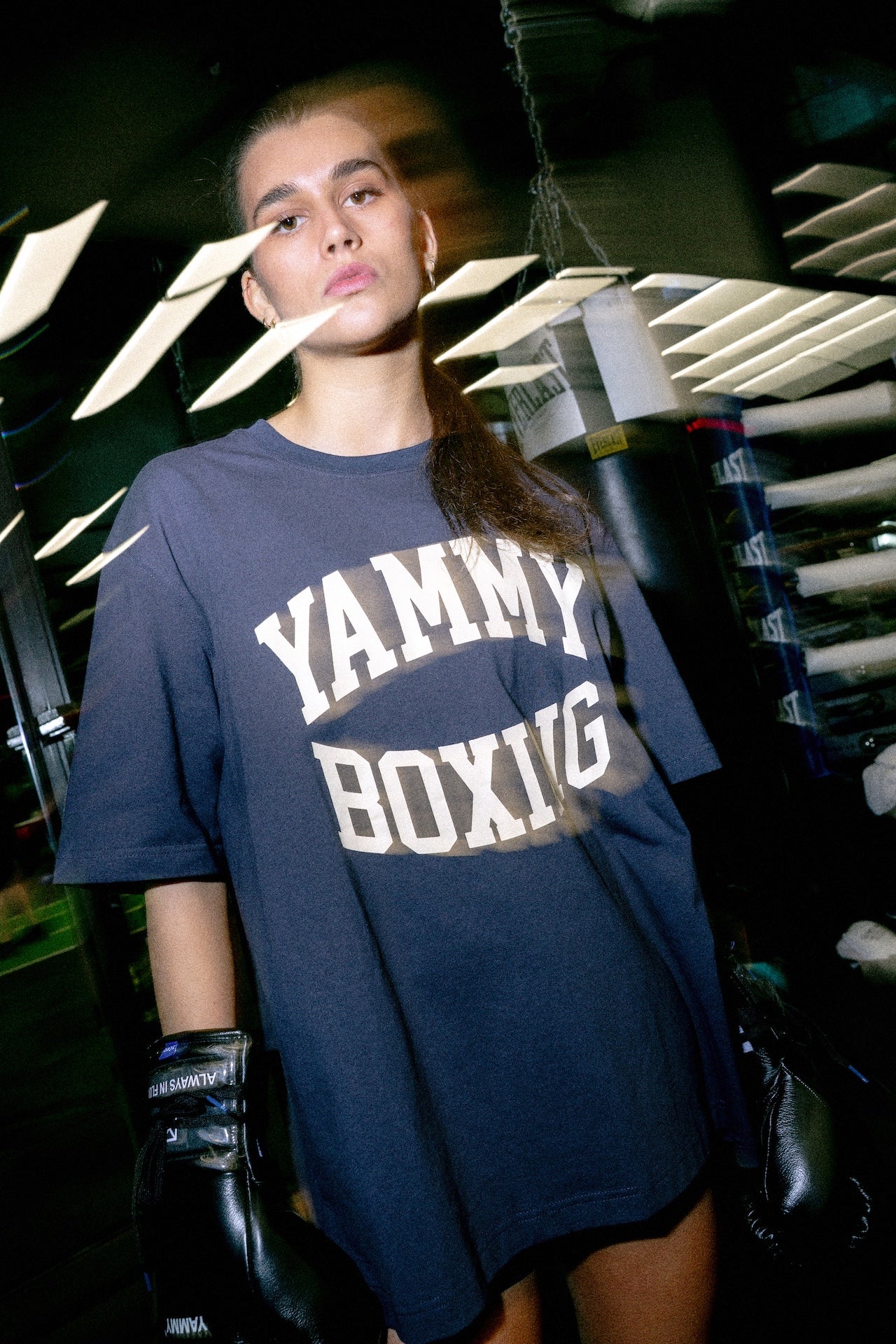 A woman in a boxing gym wearing an oversized navy blue t-shirt with 'YAMMY BOXING' printed in white varsity lettering, paired with black boxing gloves.