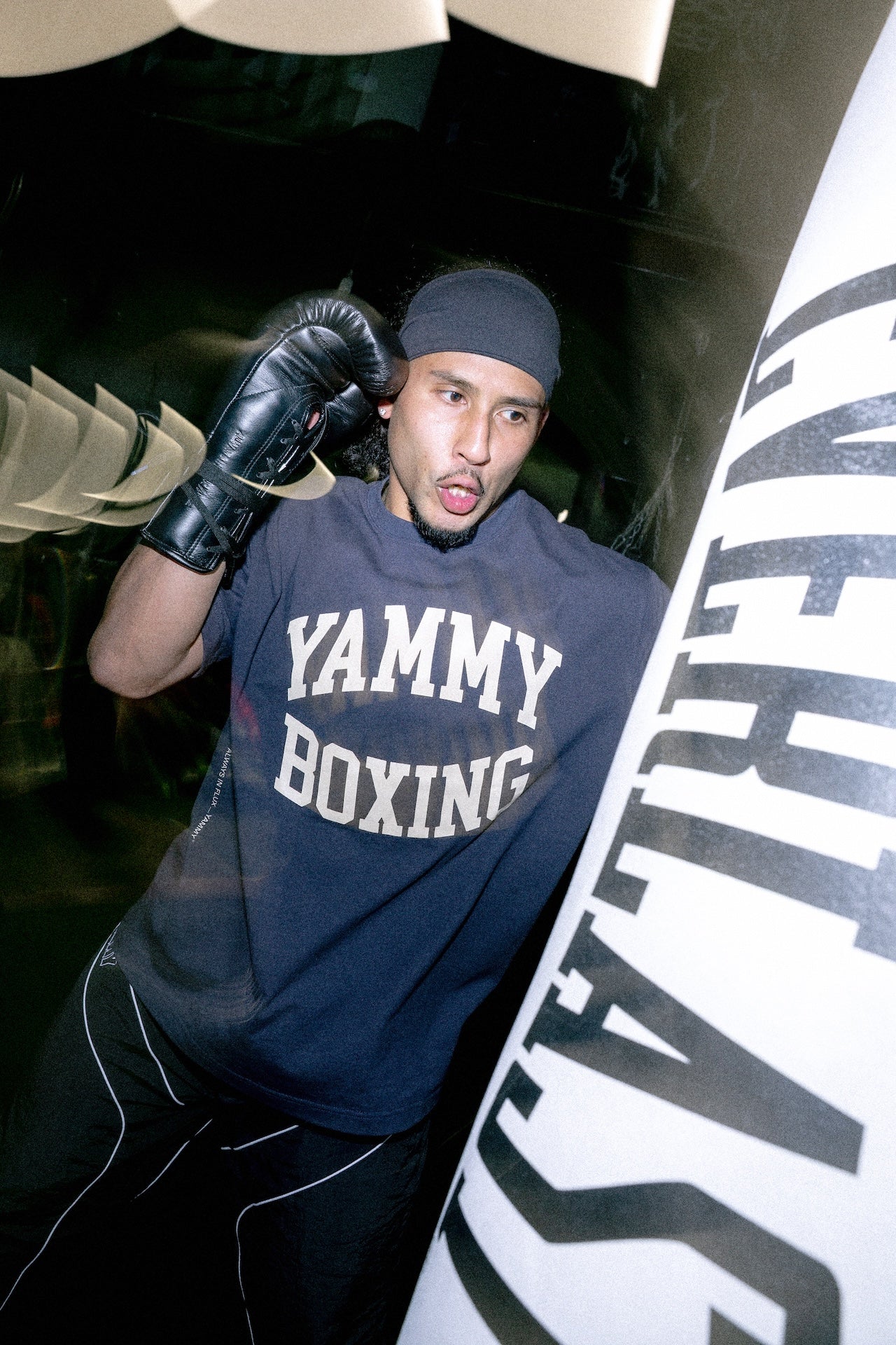 A man wearing a black beanie, black boxing gloves, and a dark gray t-shirt with 'YAMMY BOXING' printed in white, posing next to a large white and black boxing bag in a gym setting.