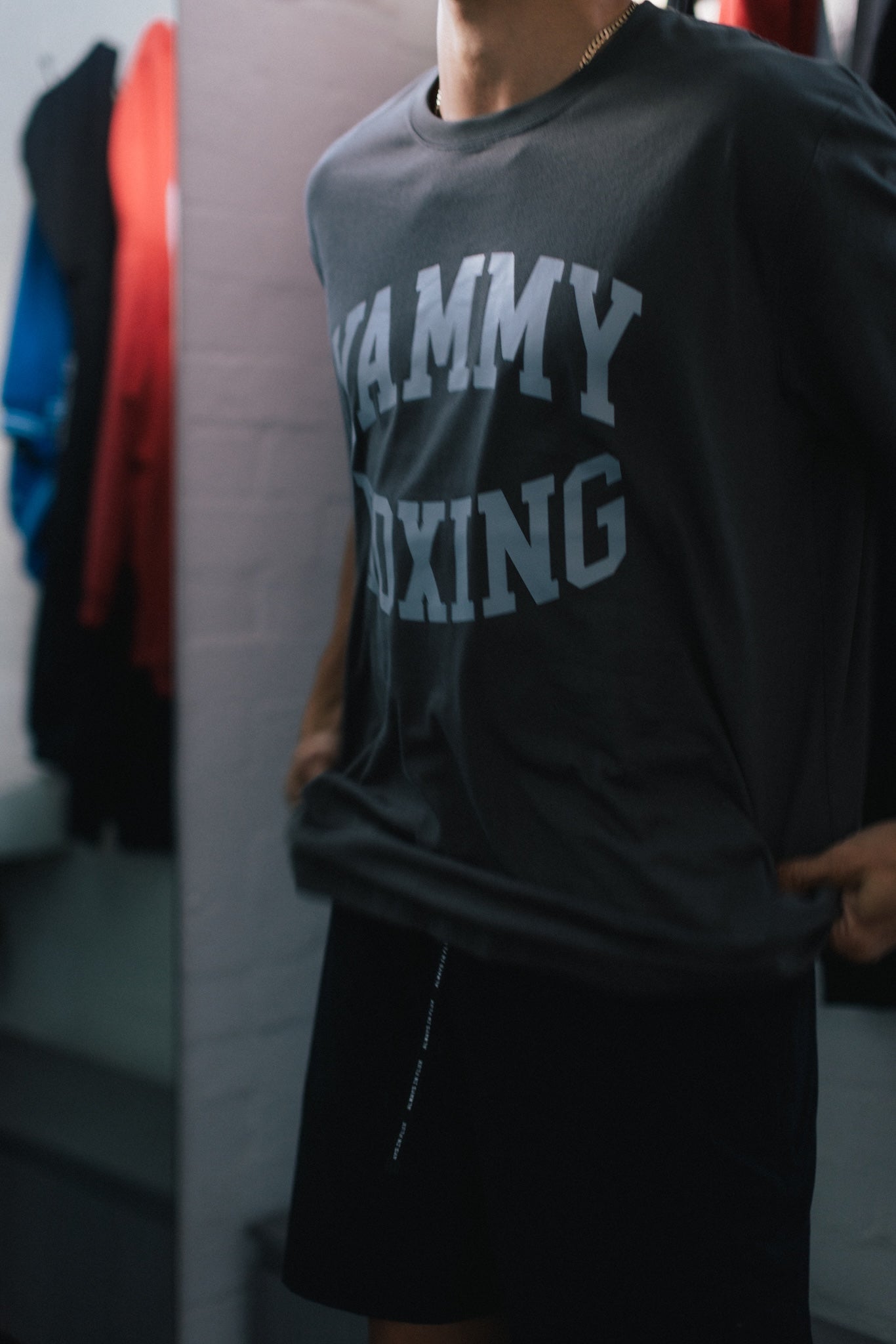 A person wearing a dark charcoal gray graphic t-shirt with 'RAMMY BOXING' printed in light gray varsity letters, paired with black shorts in what appears to be a dressing room or closet setting.