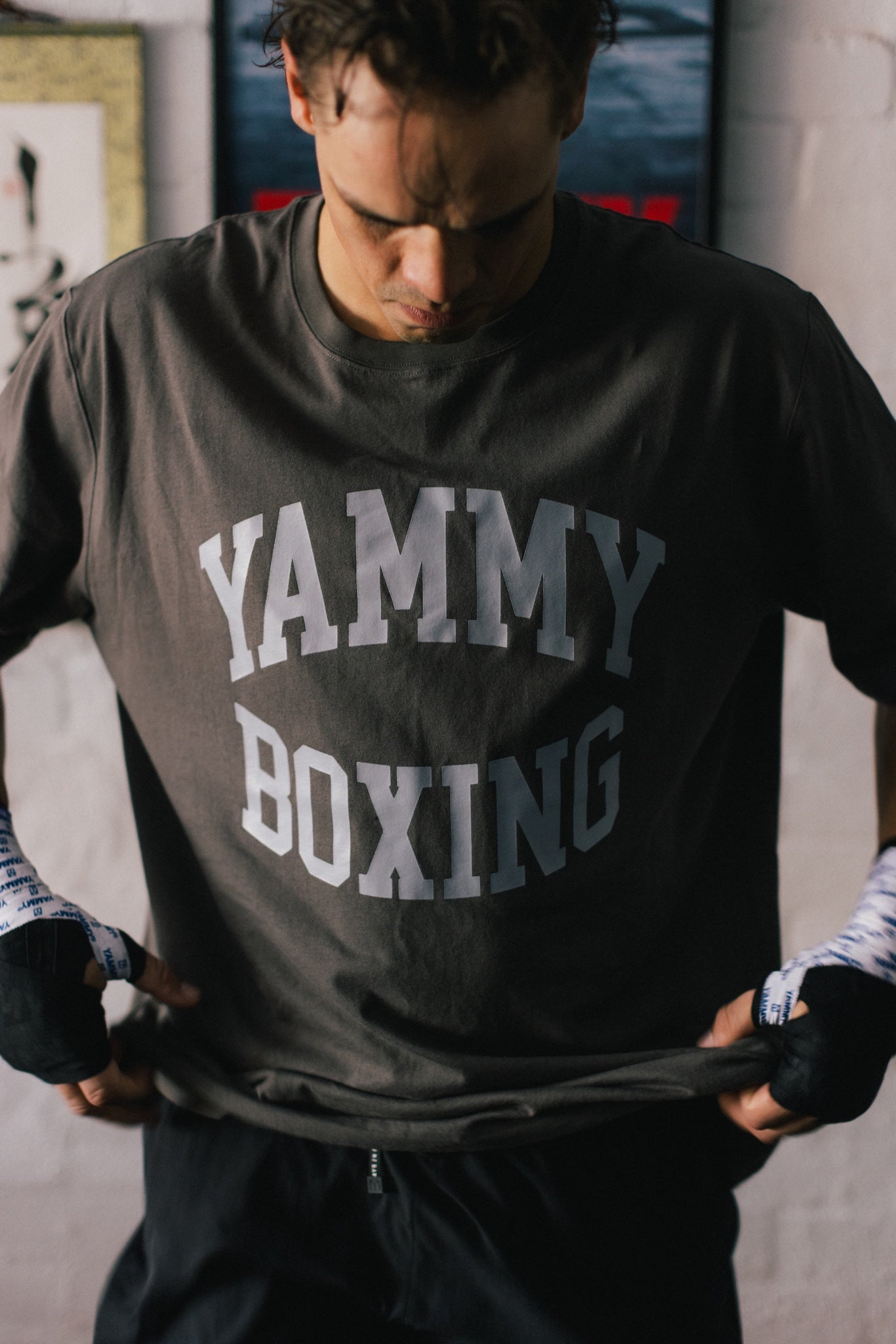 A man wearing a dark gray t-shirt with 'YAMMY BOXING' printed in white varsity-style letters, holding the hem of the shirt to show the fit.
