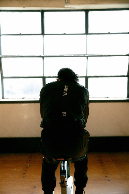 A person viewed from behind, seated on a stationary bike in a large room with a bright window. The person wears a black jacket with the brand name 'YAMMY' printed vertically on the back.