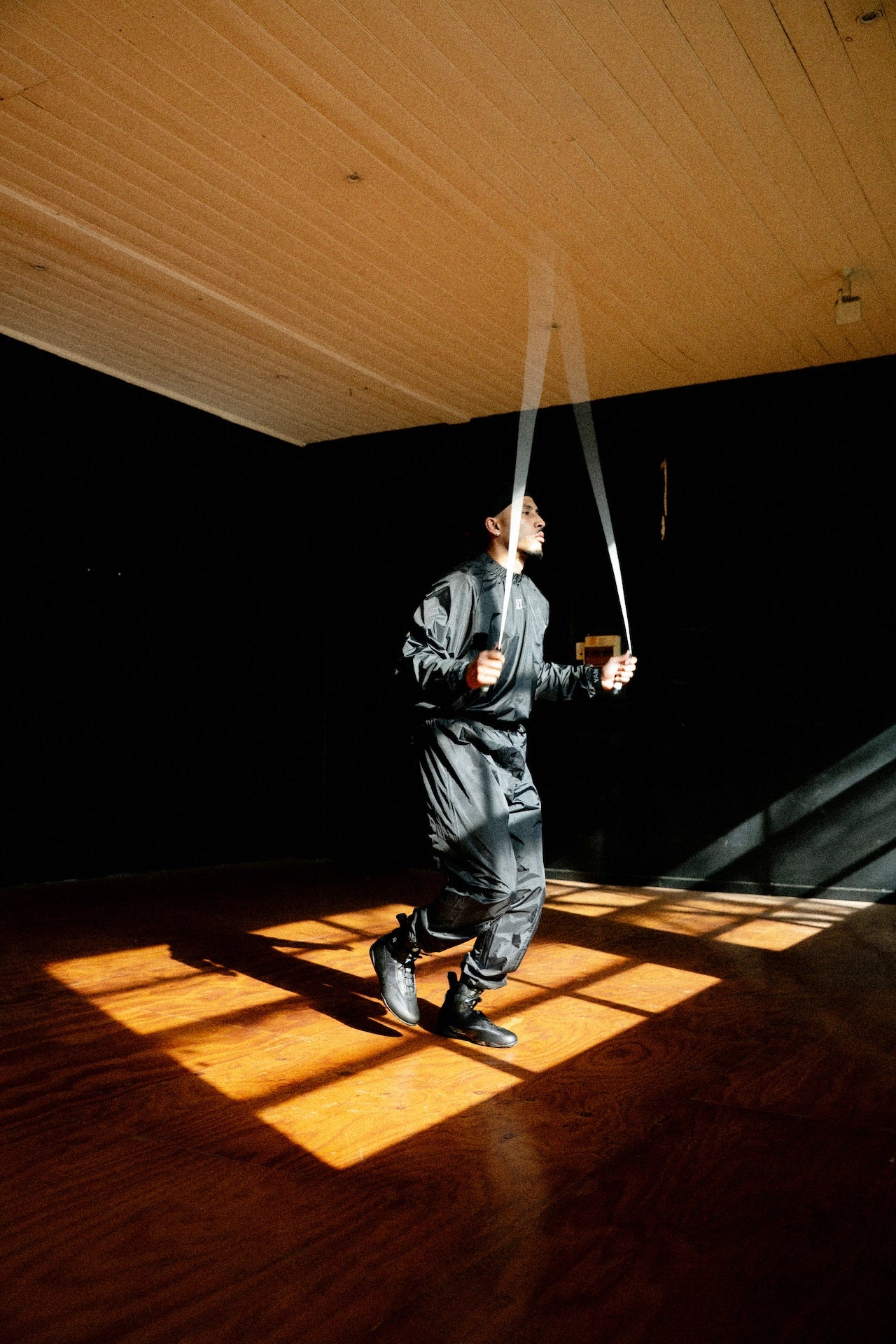 A person in a black tracksuit jumps rope in a large room with a wooden floor and ceiling, lit by dramatic sunlight and shadow.