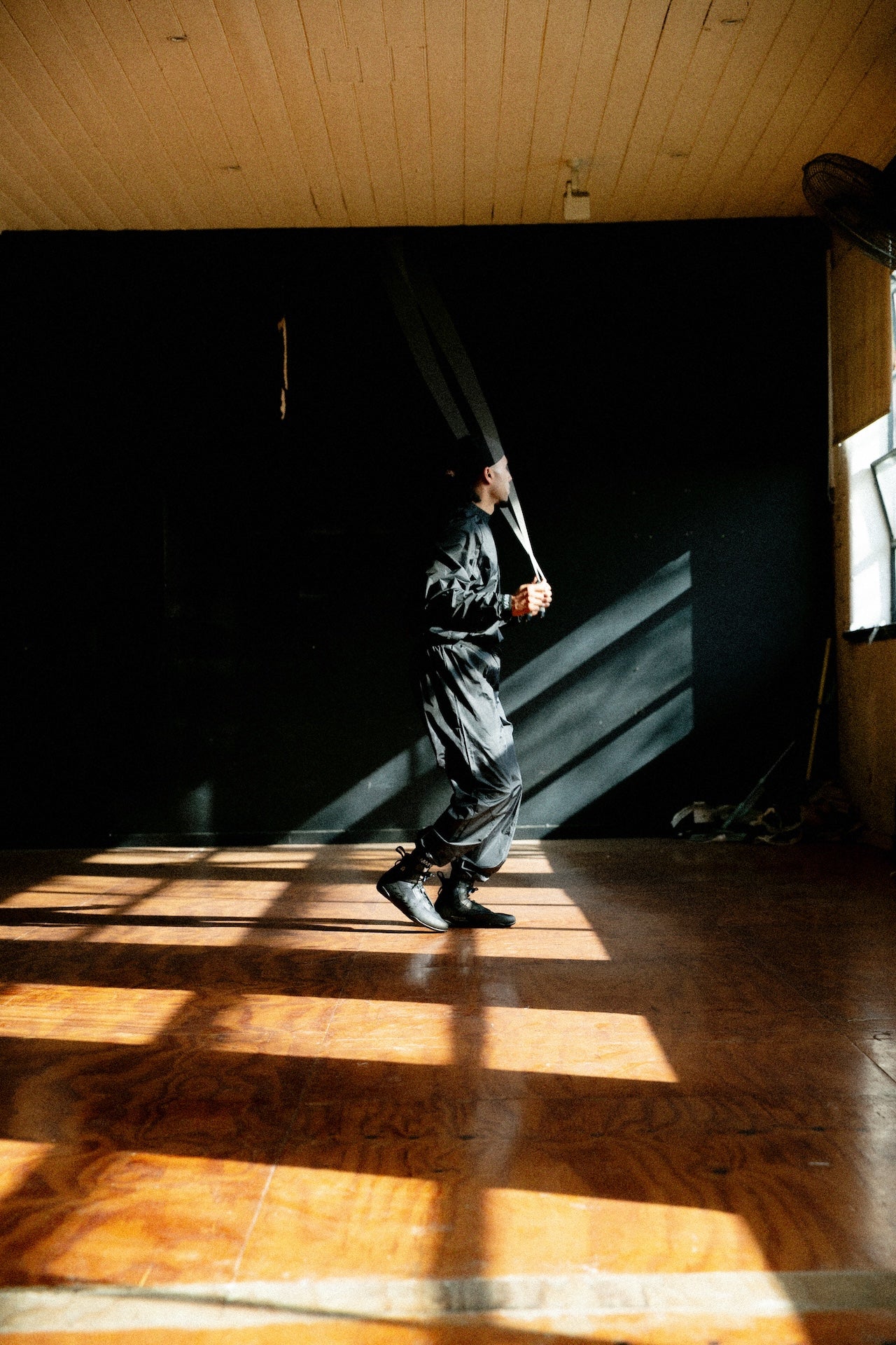A person in a black outfit walks across a wooden floor in a large room with dramatic sunlight and shadows cast across the space.
