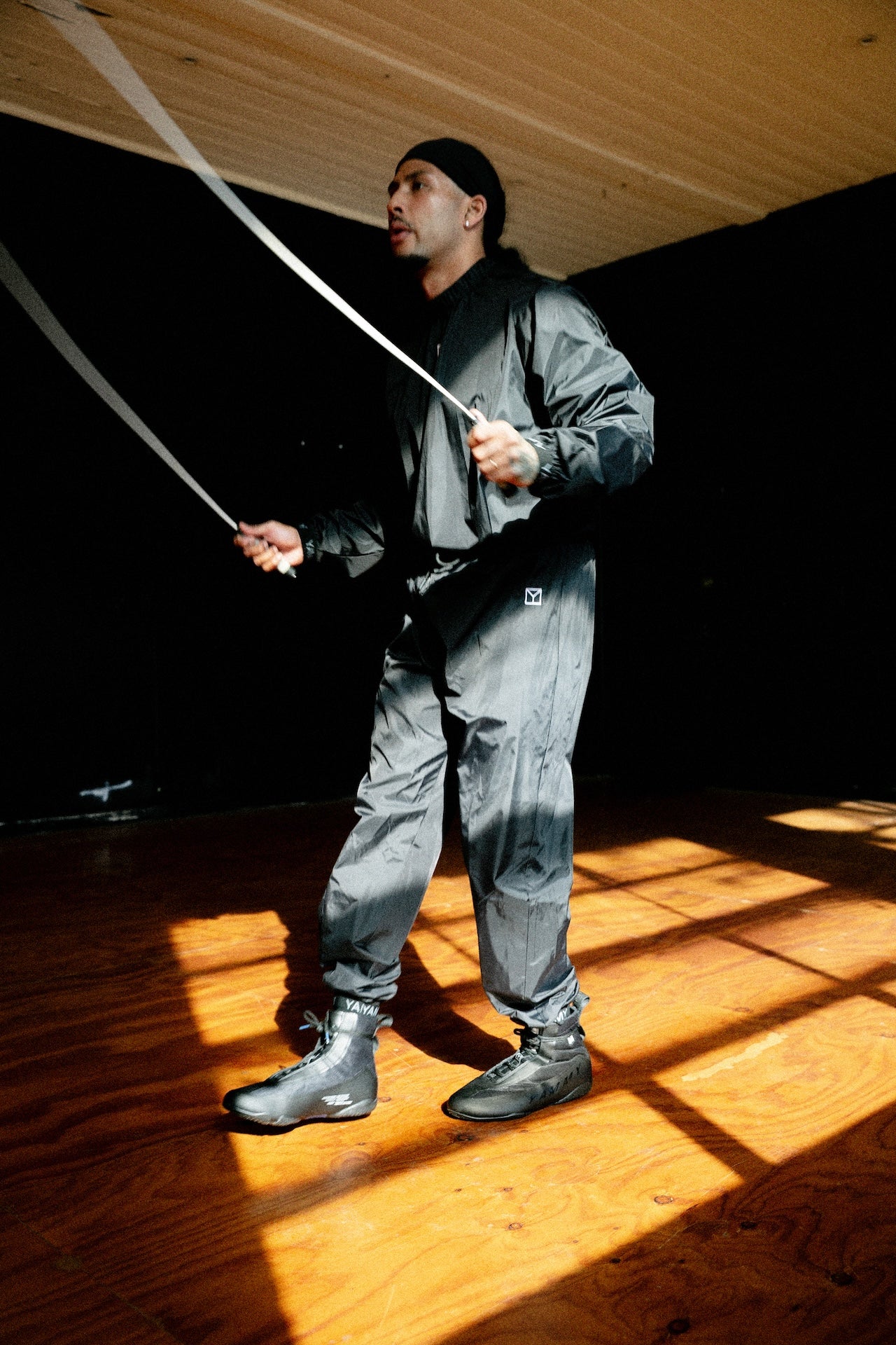 A man wearing a dark gray waterproof jacket and matching pants jumps rope in a room with a wooden floor and ceiling, lit by dramatic sunlight casting strong shadows.