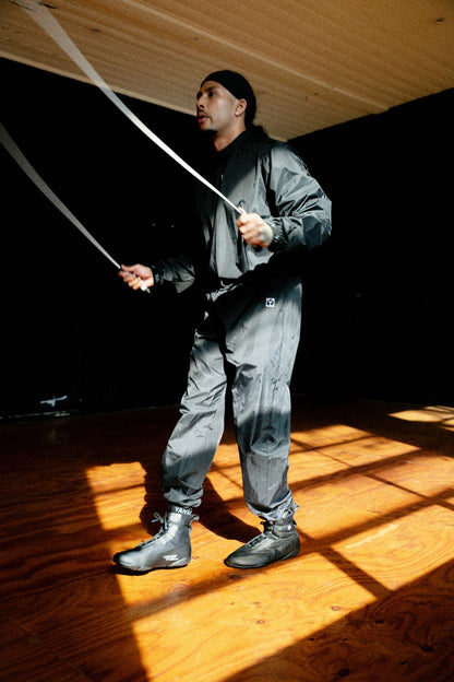 A man wearing a dark gray waterproof jacket and matching pants jumps rope in a room with a wooden floor and ceiling, lit by dramatic sunlight casting strong shadows.