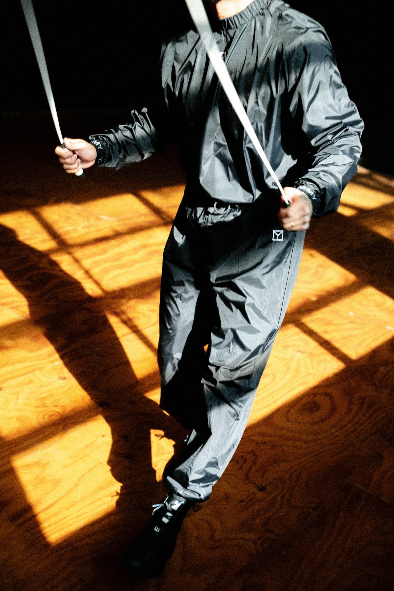 A person wearing a shiny dark gray hooded jacket and matching pants, holding a white rope or strap in a high-contrast indoor setting with strong sunlight and shadows on a wooden floor.