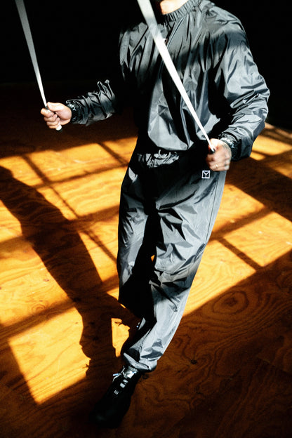 A person wearing a shiny dark gray hooded jacket and matching pants, holding a white rope or strap in a high-contrast indoor setting with strong sunlight and shadows on a wooden floor.