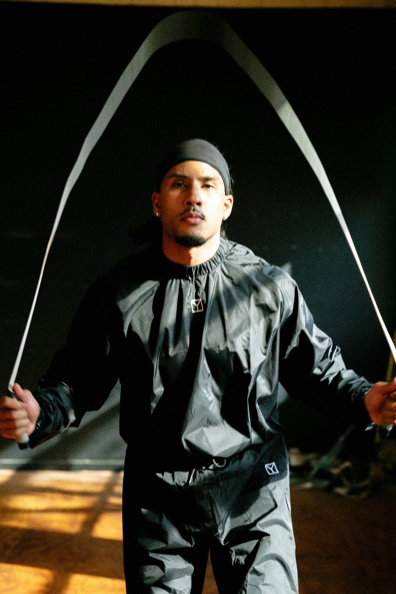 A man wearing a black athletic windbreaker jacket and matching pants jumps rope in a dark indoor setting with dramatic lighting.